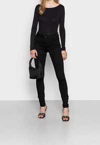 Svart långärmad bodysuit som matchas med högmidjade svarta jeans. Modellen håller en liten svart handväska och bär strapsskor i svart.