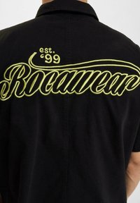 Fekete, rövid ujjú póló, nagy zöld hímzett "Rocawear" és "est. '99" felirattal a hátsó részén, sima textúrával.