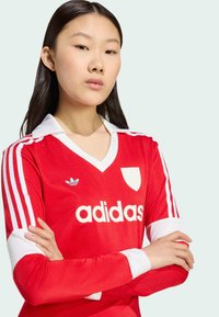 Röd långärmad fotbollsjersey med vit krage och ärmar, som har en vit "adidas"-logotyp och tre randiga detaljer, mjuk tygtextur.