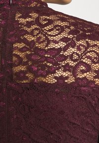 Top en dentelle bordeaux présentant des motifs floraux complexes, une texture agréable, et un détail de fermeture éclair à l'arrière. Un éclat subtil rehausse le design.