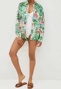 Femme portant un maillot de bain blanc, une chemise à boutons ouverte colorée avec des motifs verts et roses, des sandales à semelle brune et un collier à chaîne en argent.