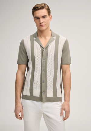 Młody mężczyzna ubrany w koszulę na krótki rękaw z pionowymi szaro-białymi paskami oraz białe spodnie, stojący na tle jednolitego tła.