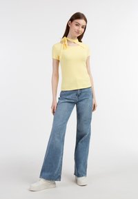 Geel geribbeld T-shirt met korte mouwen en een halsriem, gecombineerd met lichtblauwe wijde jeans en witte sneakers. Effen achtergrond.