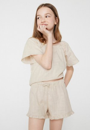 Jeune femme en haut beige à manches courtes et short assorti avec des ourlets volantés, debout, main au menton et regardant sur le côté.