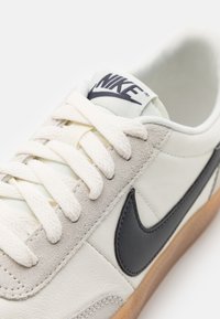 Λευκά δερμάτινα sneakers με γκρι σουέτ λεπτομέρειες, μαύρο σήμα Nike και κρεμ κορδόνια. Διαθέτει υφαντή σόλα από καουτσούκ γκοφρέ.
