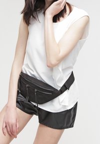 Schwarze Stoffbauchtasche mit zwei Reißverschlusstaschen, am Bauch getragen. Auch ein weißes ärmelloses Oberteil und glänzende schwarze Shorts sind sichtbar.