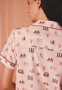 Femeie purtând o cămașă decorată cu autobuze roșii cu două etaje, câini de pază, gardieni care mărșăluiesc și cai pe un fundal roz pal.