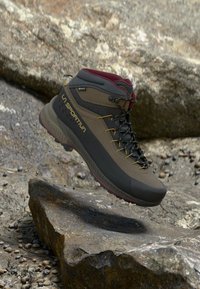 Hiking boot in dark brown and grey with maroon accents, featuring a high collar, textured sole, lacing system, and reinforced toe cap.