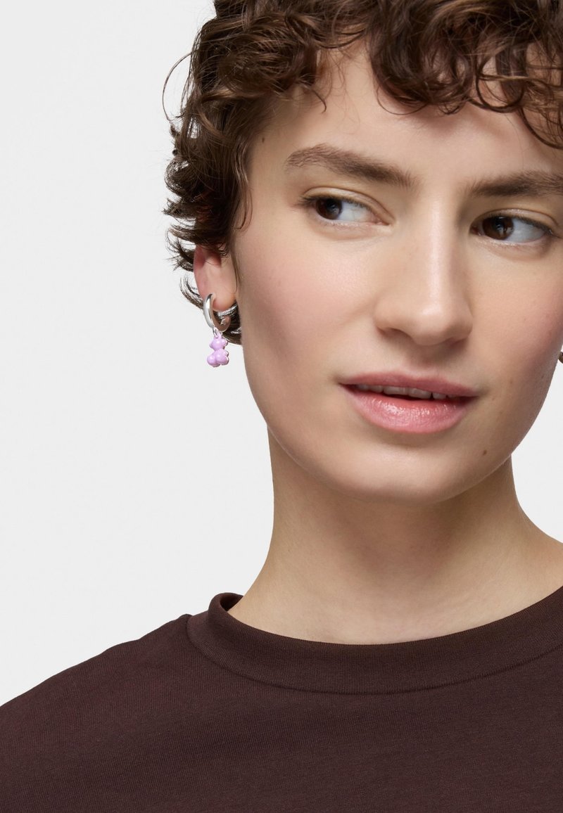 Boucle d'oreille en argent avec un pendentif en forme d'ours rose. L'arrière-plan est uni et le modèle porte une chemise marron foncé. Texture douce et finition brillante.