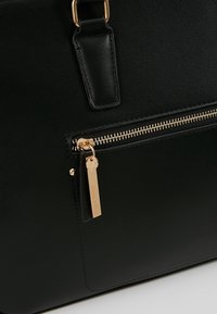 Sac en cuir noir avec fermeture éclair dorée et tirette rectangulaire dorée gravée du nom de la marque Anna Field.