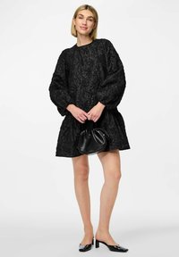 Vestido negro texturizado con mangas abullonadas y dobladillo fruncido. Combinado con un pequeño bolso negro y zapatos de tacón cuadrado de charol.