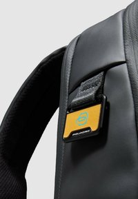 Zwarte rugzak met een getextureerde afwerking, een grijze stoffen riem en een rechthoekige logotag in geel en blauw aan de voorkant bevestigd.