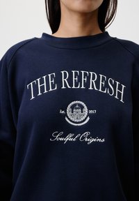 Sweat-shirt bleu marine à col rond, avec un texte brodé blanc : "THE REFRESH" et "Soulful Origins", accompagné d'un emblème central et de l'année de création.