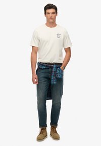 Lys beige bomulds t-shirt med et marineblåt grafisk tryk, blåternet flannel skjorte bundet om taljen, mørke jeans og tan ruskinds sko.