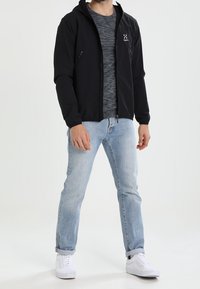 Veste noire à fermeture éclair avec capuche, dotée de deux poches zippées et d'un petit logo. Portée sur un T-shirt gris, accompagnée d'un jean bleu clair et de baskets blanches.