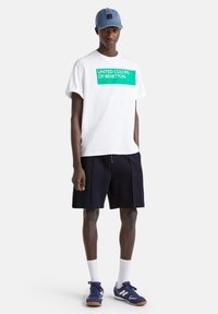 Bílý bavlněný tričko s zeleným grafickým textem, modré šortky, bílé ponožky do kotníků a modré sportovní boty. Příležitostný, uvolněný střih. Džínová kšiltovka.