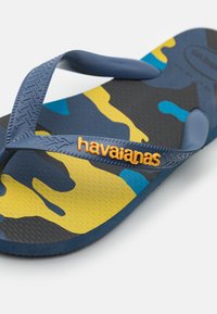 Mėlynos kamufliažo šlepetės su tekstūruota guma ir dviem plonais dirželiais. Ant dirželio yra oranžinė "havaianas" logotipo etiketė.