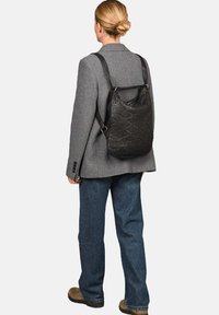 Zwarte gestructureerde rugzak met een patroon, voorzien van verstelbare schouderbanden en in driehoekige vorm, gedragen over een grijze blazer.