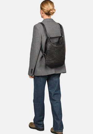 Zwarte gestructureerde rugzak met een patroon, voorzien van verstelbare schouderbanden en in driehoekige vorm, gedragen over een grijze blazer.