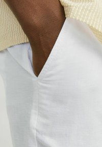 Pantalones de algodón blancos con un bolsillo lateral, combinados con una blusa a rayas amarillo claro. Las telas parecen suaves con una textura lisa.
