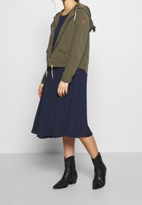 Nainen, jolla on yllä oliivinvihreä hupparitakki, tummansininen midi-mekko ja mustat nahka ankle-boots, seisoo harmaalla lattialla valkoista seinää vasten.