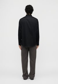 Άντρας με σγουρά μαλλιά, γυρισμένος προς τα πίσω, φοράει μακρυμάνικο μαύρο πουκάμισο και φαρδιά σκούρα γκρι παντελόνια μπροστά σε απλό ανοιχτό φόντο.