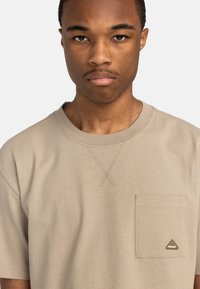 Beige Baumwoll-T-Shirt mit kurzen Ärmeln, Rundhalsausschnitt und einer Brusttasche mit einem dreieckigen Logopatch. Flache Nahtdetails am Halsausschnitt.