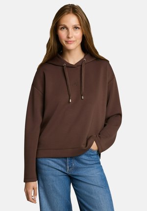Vrouw met lang lichtbruin haar, gekleed in een bruine hoodie en blauwe spijkerbroek, staand met één hand in de zak tegen een effen achtergrond.