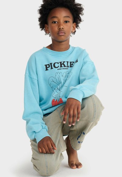 Bambino con capelli ricci in ginocchio a piedi nudi, indossa una felpa azzurro chiaro con la scritta "PICKLES" e pantaloni beige con una toppa blu.