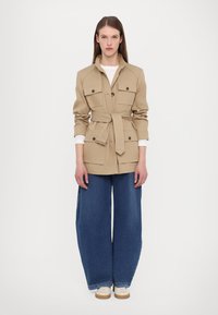 Casaco utilitário bege com cintura ajustável, grandes bolsos frontais e fecho com botão, combinado com jeans de ganga azul de perna larga e sapatos claros.