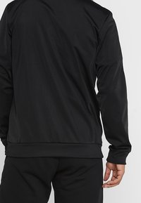 Black athletic jacket with a smooth texture, featuring long sleeves and a ribbed hem. Simple design without noticeable patterns or accents.