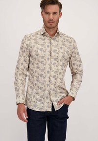 Man in een beige lange mouw overhemd met een blauw- en grijs bloemenpatroon, donkere spijkerbroek, staand tegen een effen achtergrond.