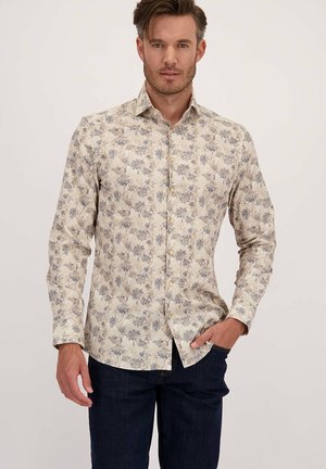 Man in een beige lange mouw overhemd met een blauw- en grijs bloemenpatroon, donkere spijkerbroek, staand tegen een effen achtergrond.