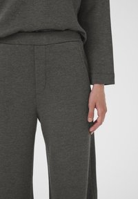 Pantaloni della tuta grigi in morbido tessuto con un design a gamba larga, dotati di una cintura liscia e tasche laterali. Primo piano dei pantaloni e di una mano.