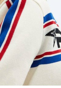 Svetr s krémovou tkaninou, červenými a modrými pruhy a černým grafickým designem. Měkká textura s neformálním, sportovním vzhledem.