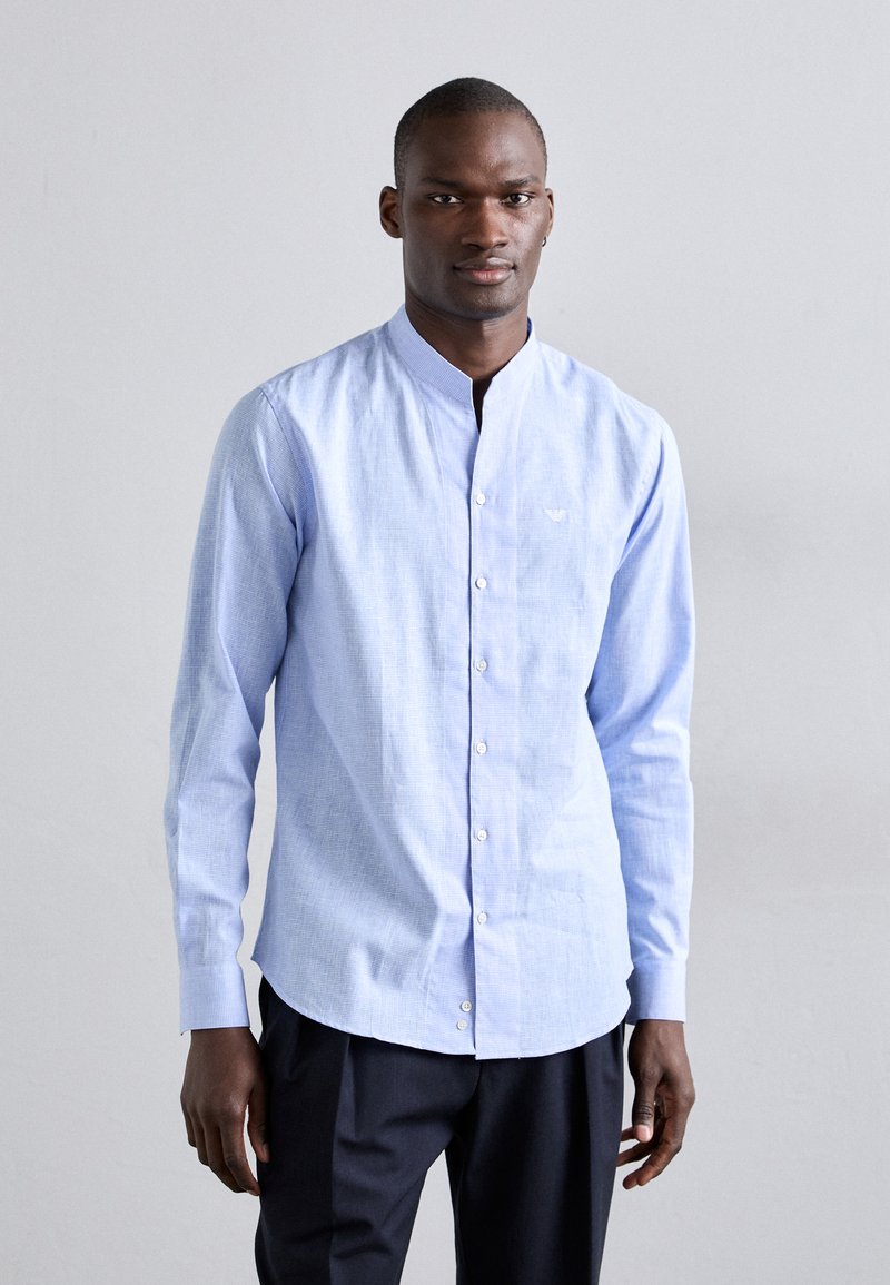 Chemise à manches longues bleu clair avec col à boutons, présentant une texture subtile et un logo sur la poitrine, assortie à un pantalon sombre.