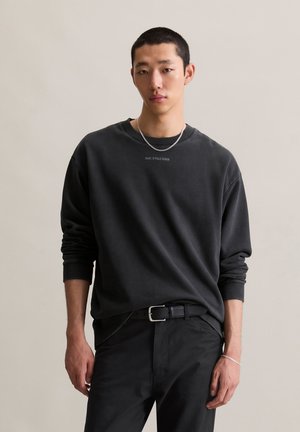 Zwarte crew-neck sweatshirt met lange mouwen, voorzien van een textuurstof en een eenvoudig logo op de borst, gecombineerd met zwarte broek en een riem.