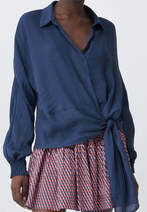 Mujer con una blusa cruzada suelta de manga larga en color azul marino y una minifalda plisada colorida con patrones geométricos en rojo, azul y blanco.