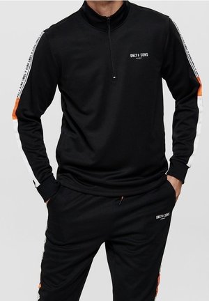 Mann trägt schwarzen Trainingsanzug mit weißen und orangen Streifen, Reißverschluss am Kragen und „ONLY & SONS“-Logo auf Brust und Hose.