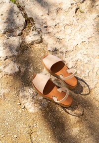 Sandales beiges avec une large bride supérieure et une bride pour les orteils, comportant une semelle intérieure marron et une semelle extérieure légère, posées sur une surface rocheuse et sablonneuse.