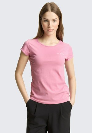 Jonge vrouw met handen in haar zakken, gekleed in een roze T-shirt met korte mouwen en een zwarte broek, tegen een effen achtergrond.