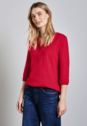 Rote, leichte Bluse mit einem geknöpften V-Ausschnitt, Dreiviertelärmeln und gerafften Manschetten, kombiniert mit dunklen Denim-Jeans.