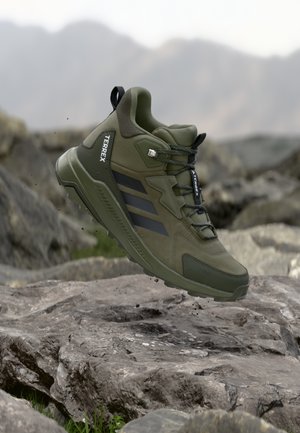 Zelená turistická obuv Adidas Terrex vo vzduchu nad skalnatým terénom s rozmazanými horami v pozadí.