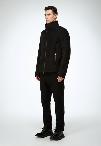 Schwarze Jacke mit Reißverschluss und hohem Kragen, ausgestattet mit seitlichen Reißverschlusstaschen und einer strukturierten Oberfläche. Kombiniert mit schwarzen Hosen und glänzenden schwarzen Schuhen.