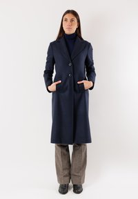 Cappotto blu in lana con vestibilità sartoriale, rever a punta, due tasche frontali e due bottoni. Abbinato a pantaloni grigi a fantasia e stivali neri.