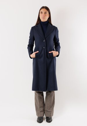 Cappotto blu in lana con vestibilità sartoriale, rever a punta, due tasche frontali e due bottoni. Abbinato a pantaloni grigi a fantasia e stivali neri.
