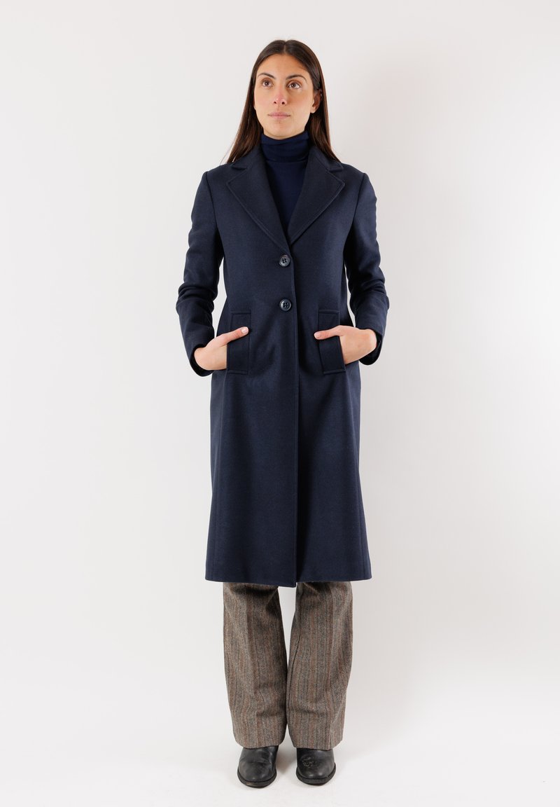 Cappotto blu in lana con vestibilità sartoriale, rever a punta, due tasche frontali e due bottoni. Abbinato a pantaloni grigi a fantasia e stivali neri.