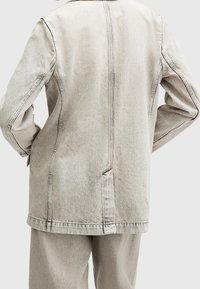 Vaaleanharmaa farkkutakki, jossa on jämäkkä muotoilu, näkyvät saumat, sivutaskut ja suora leikkaus. Minimalistinen ja tekstuuriaines.