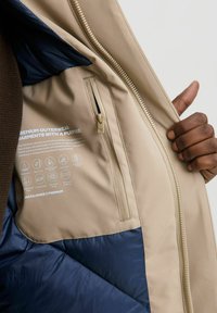 Khaki zunanja jakna z morsko modro podlogo, žepom na zadrgo, podrobnostmi o negi tkanine in teksturirano površino. Vodoodporna in izolirana zasnova.