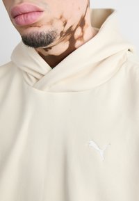 Krem dukserica s preklopljenim kragnom, izrađena od mekanog materijala. Sadrži mali bijeli izvezeni logo na prsima. Nejasna nijansa kože vidljiva.